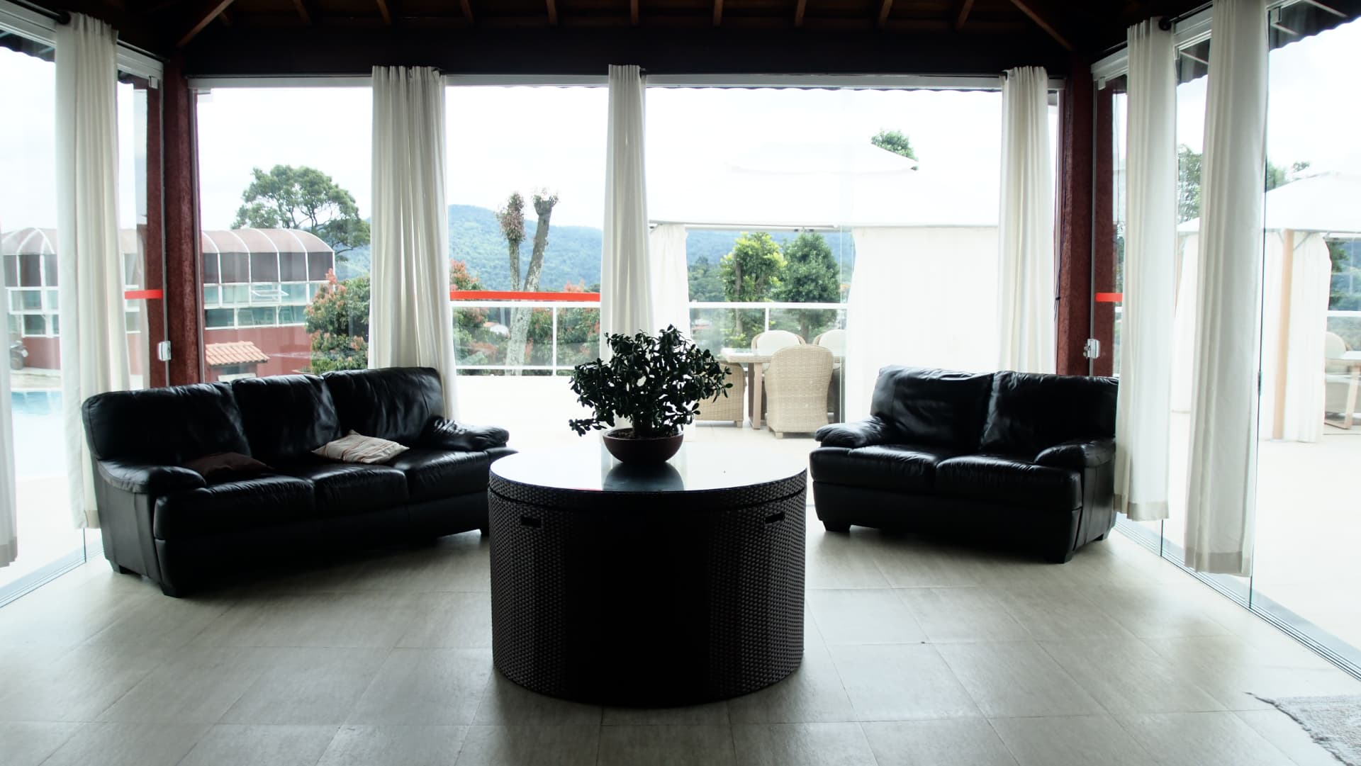 Estar interno com dois sofás pretos de couro e mesa de centro redonda, diante das paredes de vidro com vista para a natureza